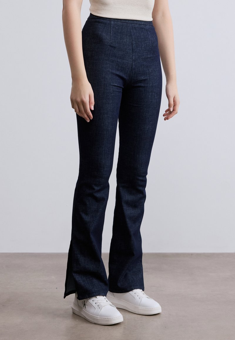 Τζιν με σκουρομπλε χρώμα, καμπάνα και ψηλή μέση, κατασκευασμένα από ντένιμ. Διαθέτουν δύο εμπρόσθιες τσέπες, ομαλή υφή και πλευρικές σχισμές στο τελείωμα.