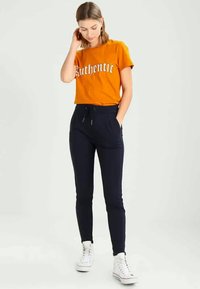 Πορτοκαλί βαμβακερή μπλούζα T-shirt με τύπωμα "Αυθεντικό", συνδυασμένη με μπλε παντελόνια jogger με πλάγιες τσέπες και κορδόνι. Λευκά αθλητικά παπούτσια.