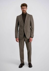 Mann steht in einem braunen Anzug mit einer Zweiknopfjacke, passenden Hosen, einem dunklen Rollkragenpullover und dunkelbrownen Lackschuhen vor einem schlichten Hintergrund.