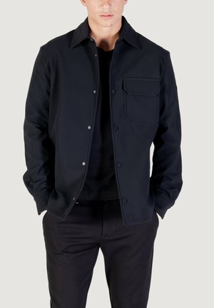 Marineblaue Jacke mit Knopfverschluss und kleiner Brusttasche, aus glattem Stoff gefertigt. Verfügt über einen Kragen und dezente Nähte. Getragen über einem schwarzen Oberteil.