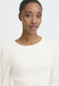 Sieviete ar tumšu ādu un matiem sasietiem atpakaļ, velkot baltu ribotu garām piedurknēm, maigi smaidot pret vienkāršu gaišu fona.