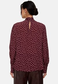 Bordeauxrote Bluse mit langen Ärmeln, hohem Kragen und Schlitzdetail am Rücken. Durchgehend mit einem cremefarbenen Sternenmuster versehen. Weicher, fließender Stoff.