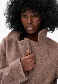 Bruine gestructureerde wollen jas met een hoge kraag en oversized mouwen. De stof lijkt zacht, wat warmte en comfort toevoegt.