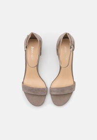 Pari taupe nubuk avattuja sandaaleja nilkkastrapeilla ja lohkokoroilla, katsottuna ylhäältä valkoista taustaa vasten.