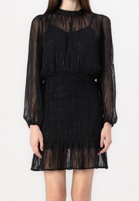 Svart klänning med genomskinliga, texturerade ärmar, med en smockad midja och en lager på lager ruffled nederkant; inkluderar subtila skimrande detaljer.