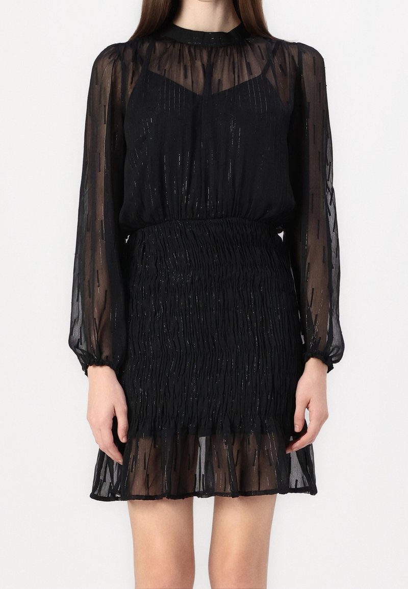 Svart klänning med genomskinliga, texturerade ärmar, med en smockad midja och en lager på lager ruffled nederkant; inkluderar subtila skimrande detaljer.