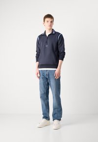 Felpa navy a mezza zip con dettagli e etichetta azzurri. Indossata sopra una maglietta bianca, abbinata a jeans blu e sneaker bianche.
