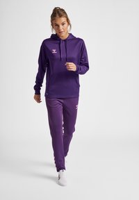 Paarse sportieve hoodie met een voorzak, een capuchon met trekkoord en een roze logo. Gecombineerd met bijpassende paarse broek met roze logo en zijaccenten.