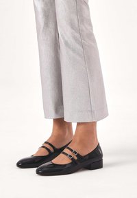 Black patent leather Mary Jane shoes with two adjustable straps and a low block heel, paired with shiny silver cropped pants.