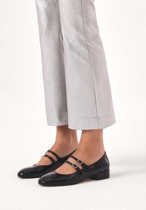 Black patent leather Mary Jane shoes with two adjustable straps and a low block heel, paired with shiny silver cropped pants.