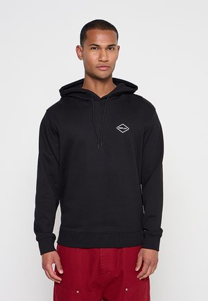 Junger Mann, der einen schwarzen Hoodie mit kleinem "Replay"-Logo auf der Brust und roten Hosen trägt, steht vor einem einfarbigen grauen Hintergrund.