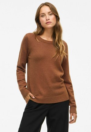 Femme aux cheveux longs et ondulés portant un pull marron à manches longues et un pantalon noir, se tenant avec une main dans la poche devant un fond uni.