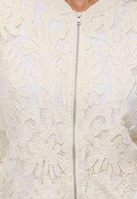 Gros plan sur un tissu en dentelle crème avec des motifs floraux et une fermeture éclair argentée au centre, porté sur un vêtement blanc.