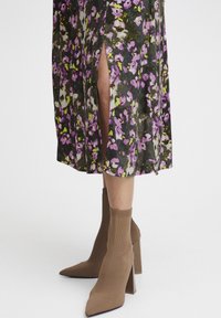 Robe fleurie avec des motifs violets, verts et blancs ; présente des fentes sur les côtés. Portée avec des bottines à chaussette beige, à bout pointu et à talon épais.