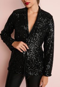 Chaqueta de lentejuelas negras con escote en V profundo, mangas largas y forma ajustada. Textura brillante con acentos metálicos. Combinada con leggings negros.