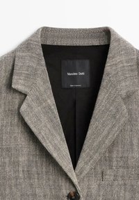 Grå sildeben blazer med reverskrave, enkelt knaplukning og sort indvendig label. Tekstureret stof med subtilt mønster.