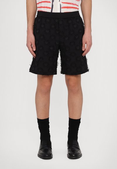 Shorts noirs avec des appliqués floraux texturés, taille élastique, coupe mi-genou et design à l'avant plat. Portés avec des chaussettes noires et des chaussures.
