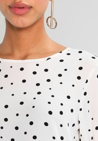 Blusa bianca con pois neri, texture liscia, scollo rotondo, tessuto trasparente, maniche corte, abbinata a un orecchino circolare dorato.