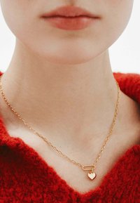 Collar de cadena de oro con un diseño de eslabones ovalados y un pequeño colgante en forma de corazón. Llevado sobre un suéter rojo texturizado.