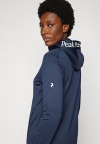 Tamsi mėlyna gobtuvo džemperis su baltu "Peak Performance" logotipu ant gobtuvo ir logotipo detale ant rankovės. Sklandžiama audinio tekstūra.