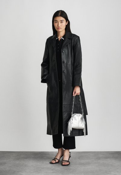 Trench coat nero in pelle con chiusura a bottoni, maniche lunghe e tasche laterali. Abbinato a una borsa argentata e sandali neri con cinturini.