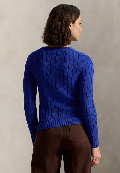 Flettet genser i livlig blåfarge, med rund hals og ribbestrikkede mansjetter. Brukt med brune bukser, som viser en tettsittende silhuett.