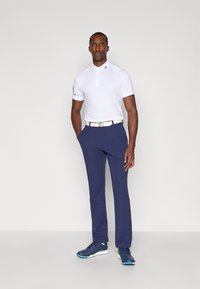 Hvit polo-skjorte med kort erme og krage. Navy blå bukser. Bæreren har et lyst belte og blå treningssko. Enkelt og rent design.