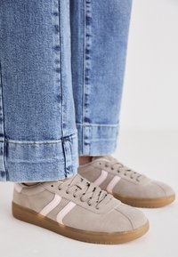 Baskets en suède gris avec des accents roses, lacets gris clair et semelles en caoutchouc beige. Un jean à jambe large avec des ourlets est visible.