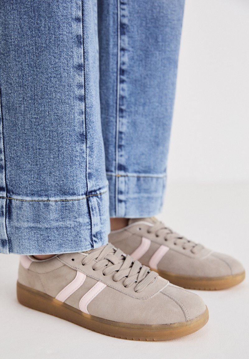 Baskets en suède gris avec des accents roses, lacets gris clair et semelles en caoutchouc beige. Un jean à jambe large avec des ourlets est visible.
