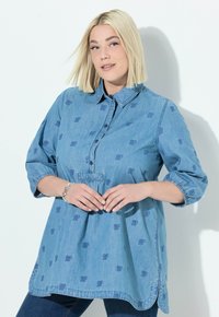 Camicia di jeans azzurro chiaro con bottoni sul davanti, maniche corte e delicati motivi a farfalla. Presenta un orlo arcuato e tasche.