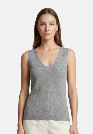 Donna con capelli lunghi e castani che indossa un top grigio a maglia senza maniche con scollo a V e pantaloni chiari su uno sfondo bianco uniforme.
