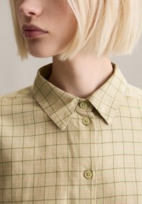 Camicia verde chiaro a quadri con colletto abbottonato, caratterizzata da un motivo a griglia con accenti rosa e due bottoni rotondi al collo.