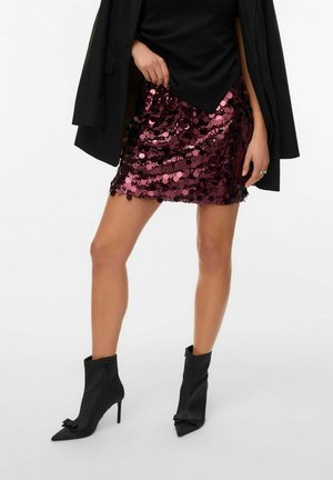 Jupe courte ajustée en bordeaux avec de grandes paillettes. Associée à des bottines noires à bout pointu et avec des accents de nœud.