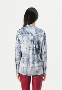 Camicia a maniche lunghe in tessuto azzurro chiaro con stampa tropicale di palme. Presenta un colletto, polsini con bottoni e un orlo curvo. In contrasto con pantaloni rossi.