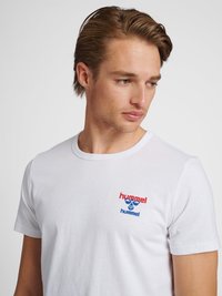 Młody mężczyzna z jasnobrązowymi włosami, noszący białą koszulkę z czerwono-niebieskim logo "hummel", patrzy w dół w lewo z neutralnym wyrazem twarzy.