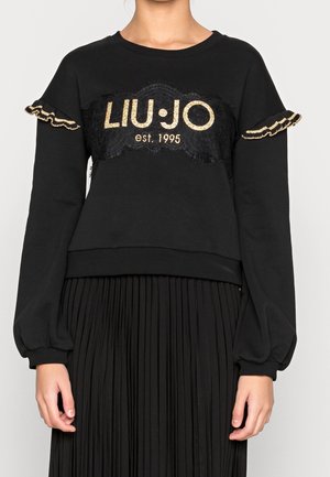 Femme portant un sweat-shirt noir avec le texte doré "LIU·JO est. 1995" et des manches à volants, assorti à une jupe noire plissée.