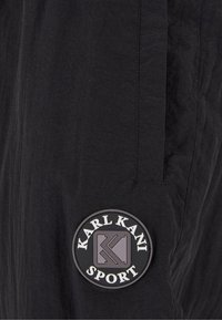 Pantalones deportivos negros con tela texturizada. Presentan un parche redondo de goma con el texto "KARL KANI SPORT" en el lado izquierdo.