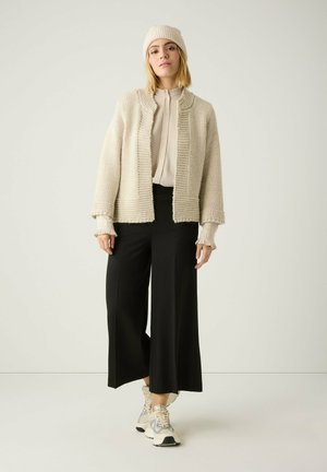 Woman wearing a cream knit beanie and cardigan over a beige blouse, black wide-leg pants, and white and silver sneakers, standing against a plain wall.