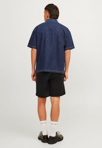 Denim short-sleeve shirt in dark blue, featuring a relaxed fit, open collar, and contrasting stitching, paired with black shorts and footwear.
