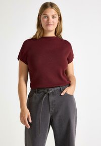 Vínová halenka s krátkým rukávem vyrobená z měkk�é tkaniny s vysokým výstřihem, v kombinaci s vysokými šedými kalhotami s páskem na uvázání.