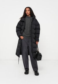 Casaco puffer preto com silhueta longa, apresentando um quilt horizontal largo, fecho com zipper e um corte descontraído, usado sobre um suéter cinza e calças.