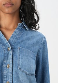 Gros plan d’une personne portant une chemise en denim bleue à boutons avec une poche poitrine, montrant le bas du visage et des cheveux bouclés foncés.
