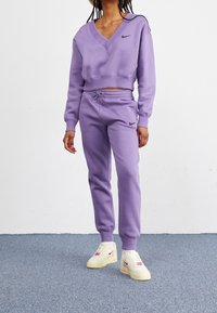 Kvinna klädd i en lila Nike cropped V-ringad sweatshirt och matchande joggers med krämfärgade Nike sneakers, stående på en blå matta.