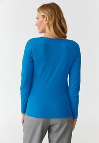 Langarmshirt in lebhaftem Blau, aus glattem Stoff gefertigt, mit Rundhalsausschnitt und taillierter Passform, kombiniert mit grauen Hosen.