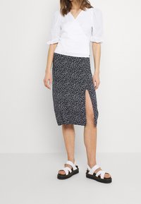 Haut blanc à manches bouffantes avec un col en V, associé à une jupe midi bleu marine à imprimé floral avec une fente latérale, et des sandales blanches à brides.