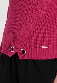 Fuchsia gestrickter Pullover mit geflochtenen Details und silbernen Ösen über schwarzen Hosen, mit einem kleinen Metall-Logo an dem Saum.