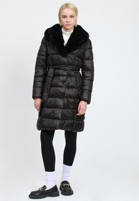 Donna che indossa un cappotto trapuntato nero cintrato con un ampio colletto in pelliccia sintetica, leggings neri, calze bianche e mocassini neri con dettagli dorati.
