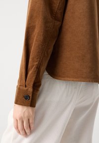 Chemise en velours côtelé marron à manches longues avec des poignets à boutons noirs, portée par-dessus un pantalon blanc à côtes verticales, main reposant sur le côté.