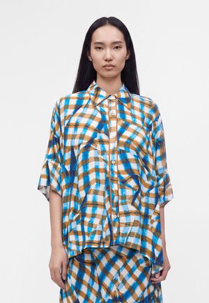Femme aux longs cheveux noirs portant une chemise ample boutonnée et une jupe avec un motif à carreaux bleu, marron et blanc très marqué sur un fond uni.