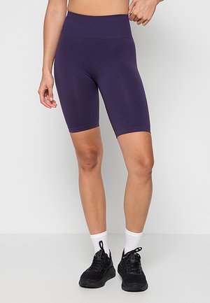 Personne portant un short de vélo violet taille haute, des chaussettes blanches et des chaussures de sport noires, debout devant un fond uni.